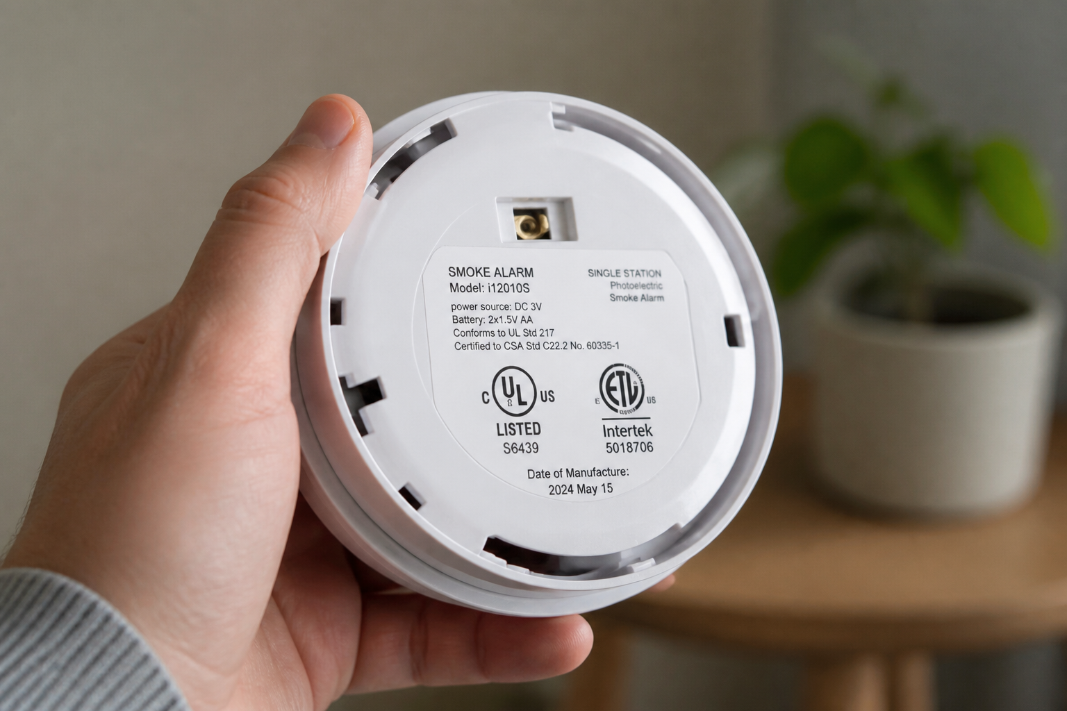 The back of a white smoke alarm held in one hand, showing the printed certification mark, model number and date of manufacture label