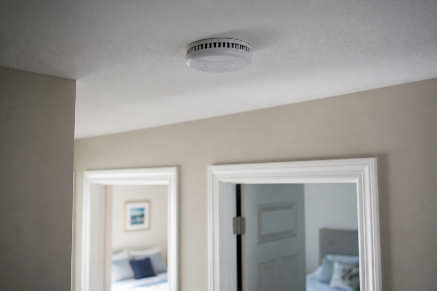 White ceiling mounted smoke alarm installed in a hallway several inches clear of the wall, with two bedroom doors visible behind it